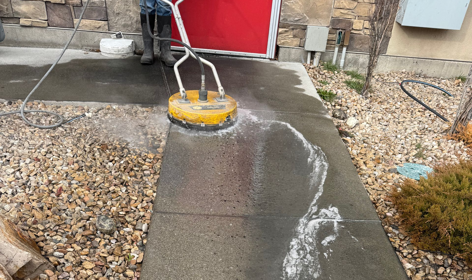 A person uses a yellow rotary surface cleaner to pressure wash a concrete walkway next to a gravel area.