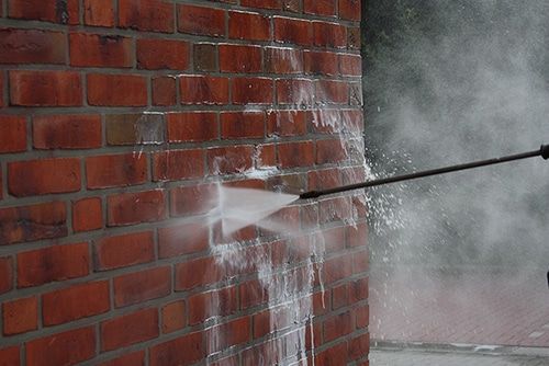 A pressure washer sprays a focused stream of water against a red brick wall, cleaning the surface.
