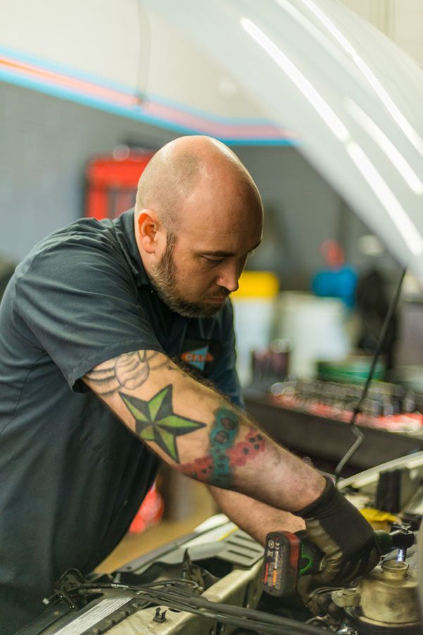 A man with tattoos is working under the hood of a car. | The Garage in Renton