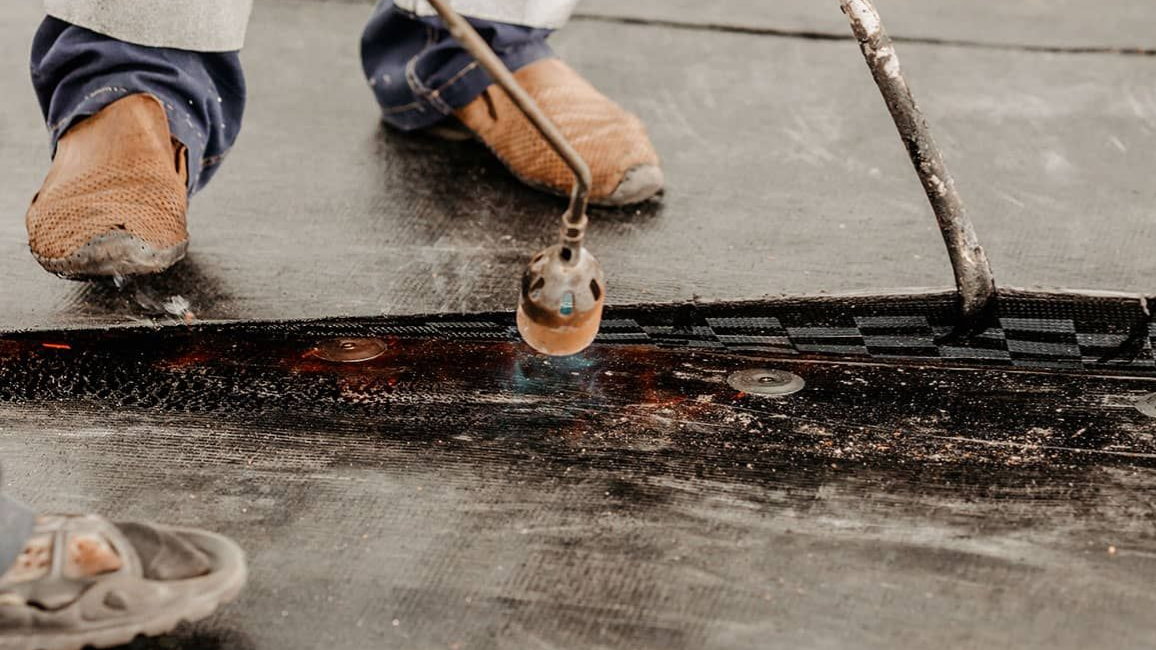 Person using a torch to seal seams on a black roofing surface.