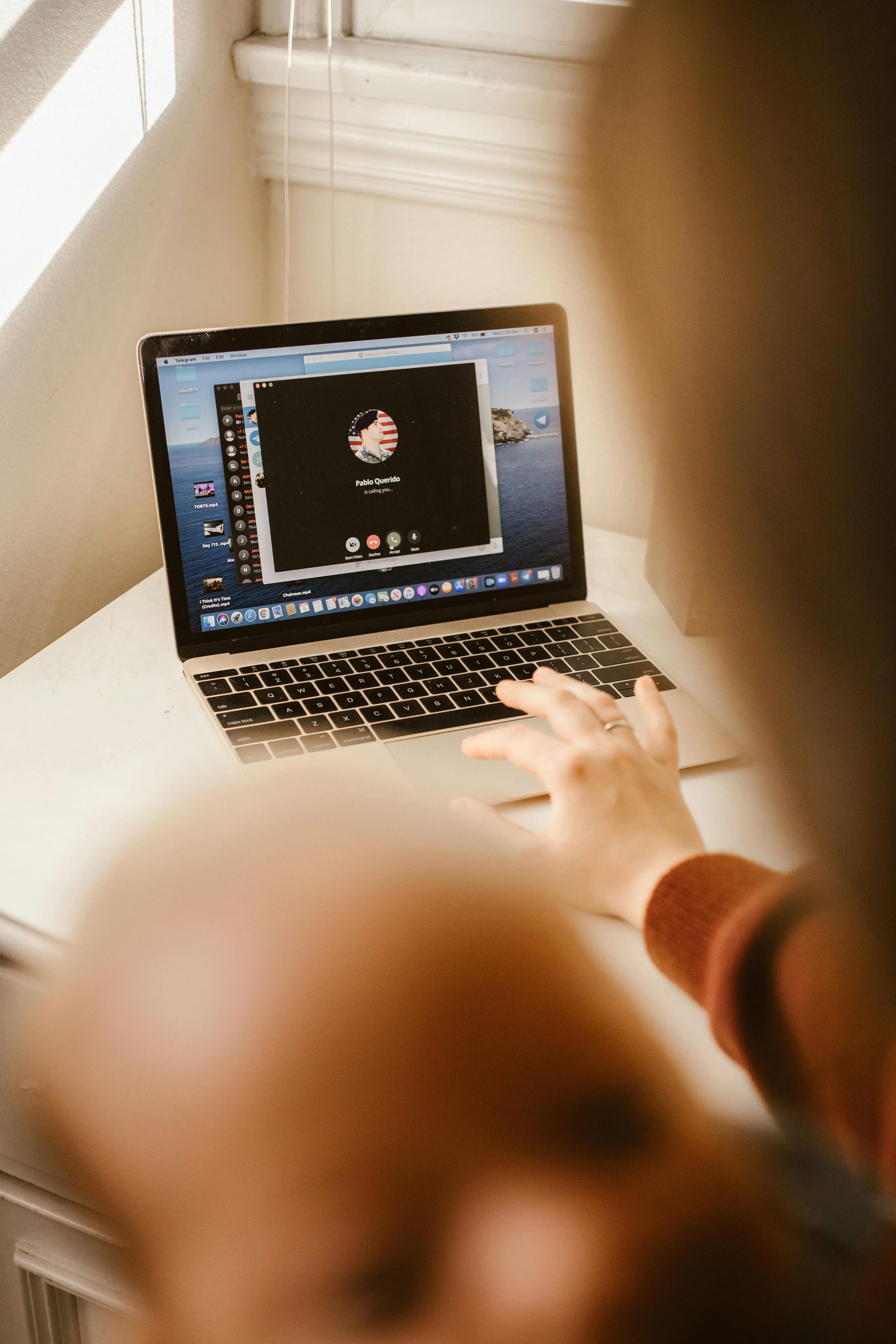 A person’s hand rests on a laptop keyboard while participating in a video call on a desk near a window.