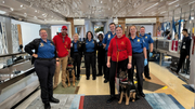 A group of security personnel in blue uniforms and red sweatshirts stand with two working dogs in an airport checkpoint.