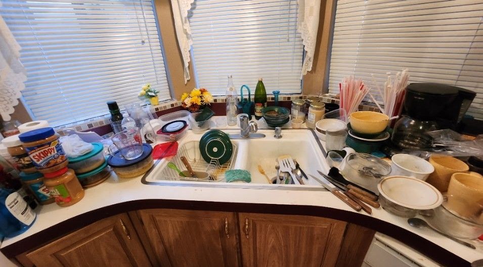 Cluttered kitchen counter with sink full of dirty dishes. Various containers and items on the countertop.