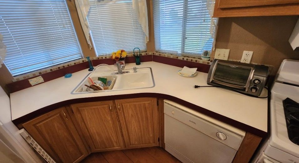 Kitchen countertop with sink, dishwasher, and toaster oven. Wooden cabinets below, blinds above.