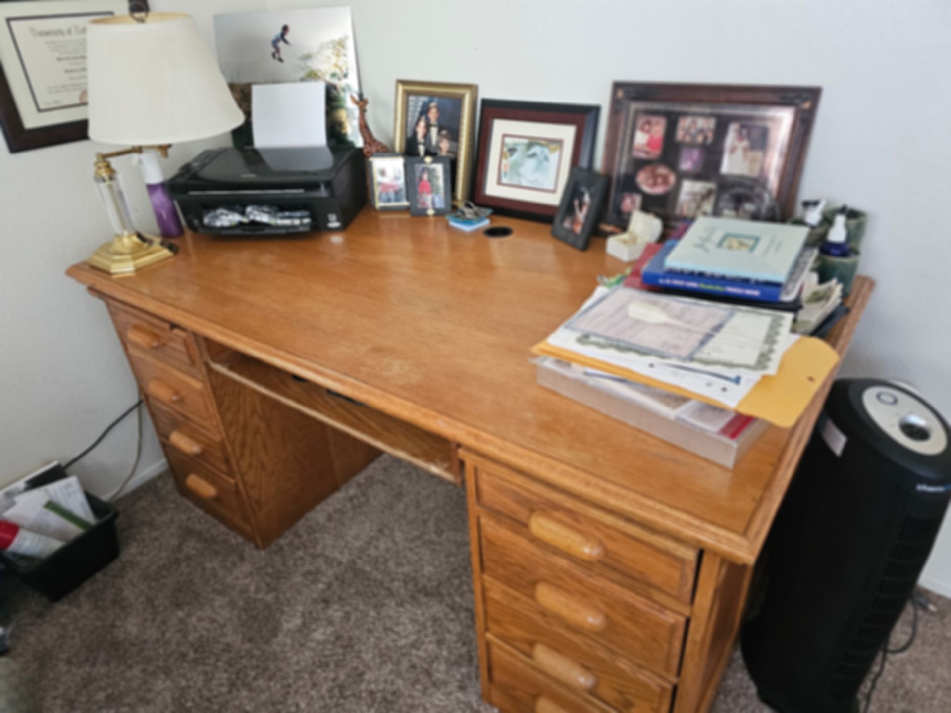 Wooden desk with drawers, lamp, printer, framed photos, and documents in a corner room.