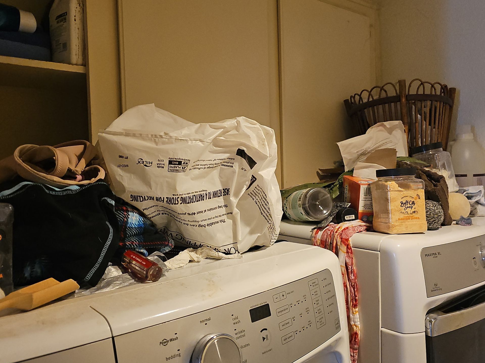 Laundry room with cluttered washing machine top.  Items include laundry bag, clothes, supplies, and basket.