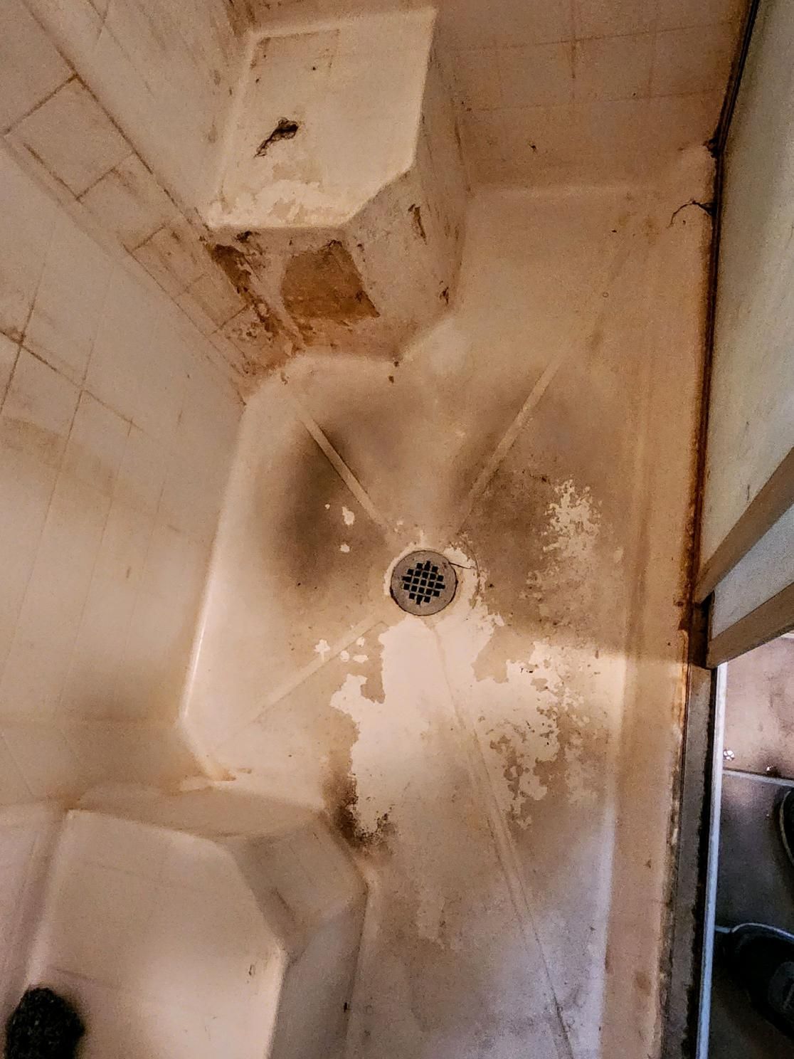 Dirty, stained shower stall with central drain; tan and brown discoloration, peeling paint.