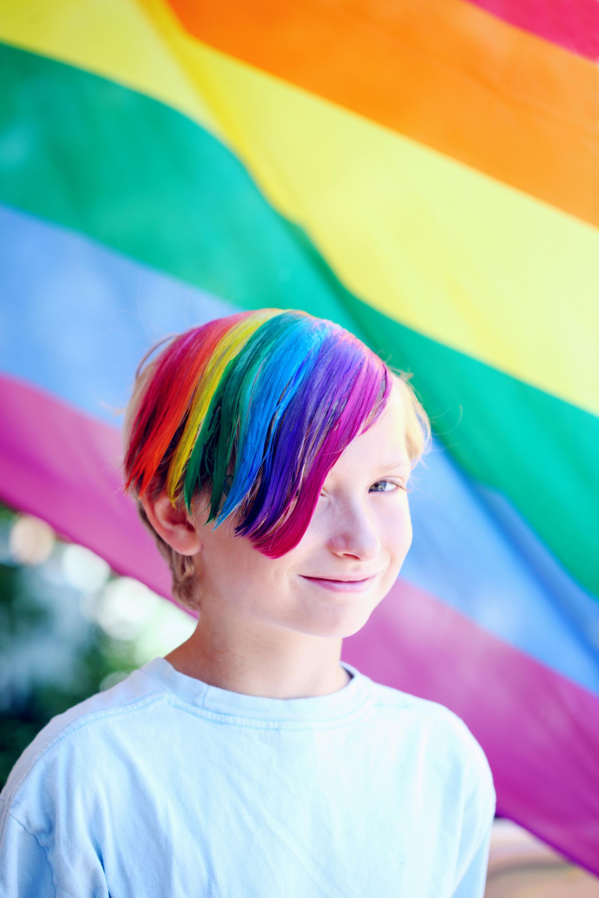 A young boy with rainbow hair is standing in front of a rainbow flag.