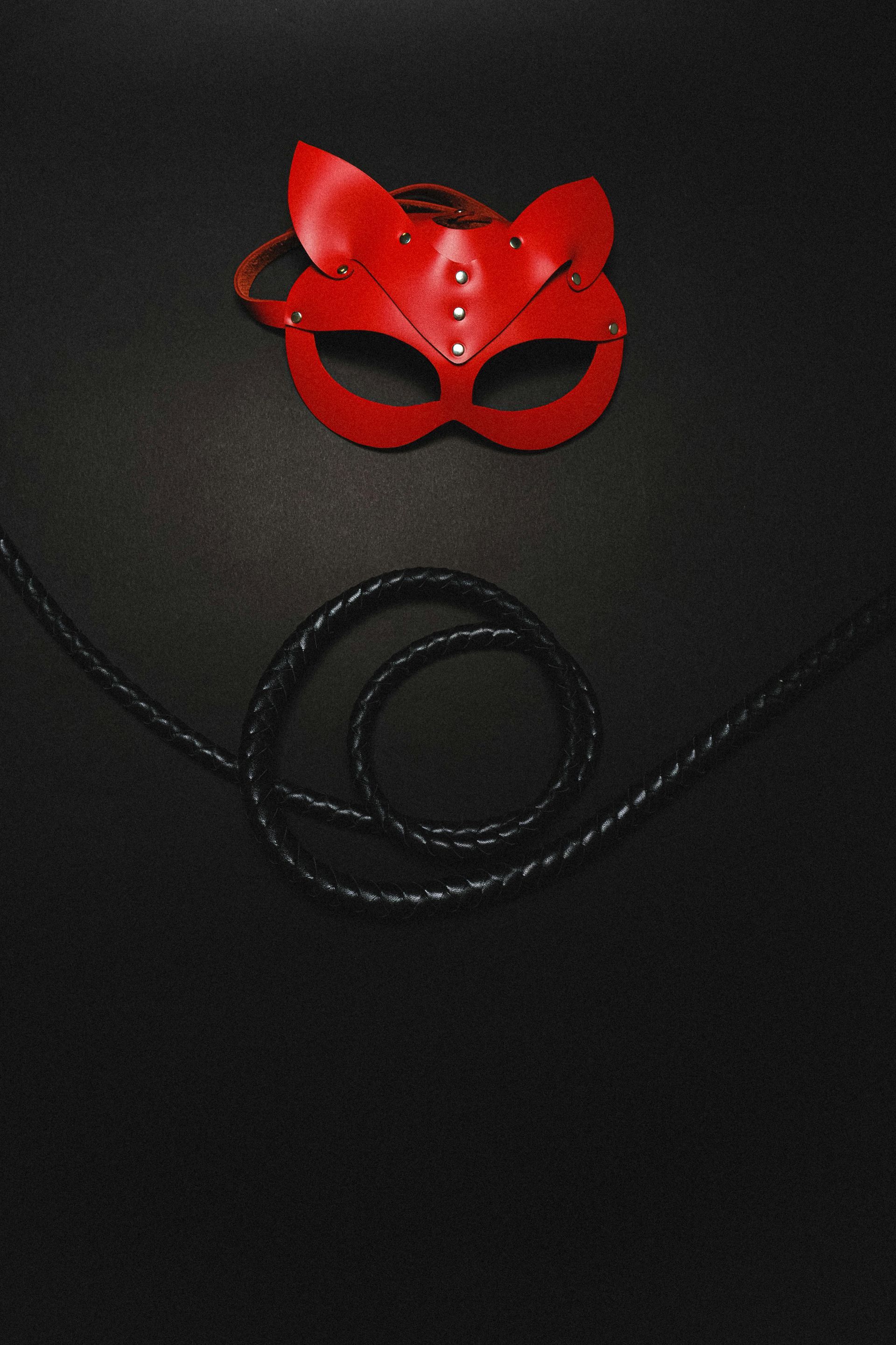 A red cat mask and a black whip are on a black surface.