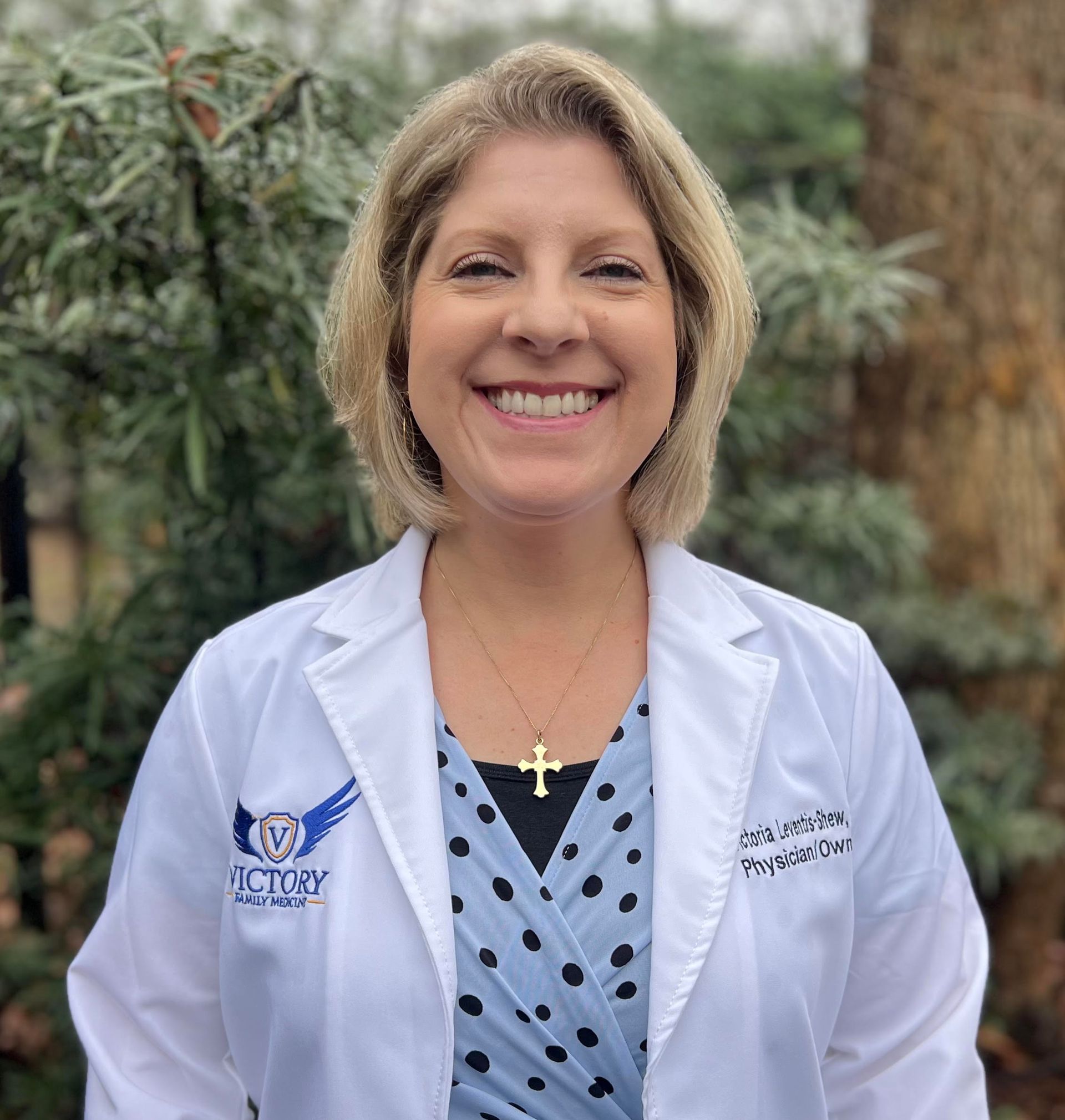 A woman in a white lab coat and blue scrubs with a stethoscope around her neck is smiling.