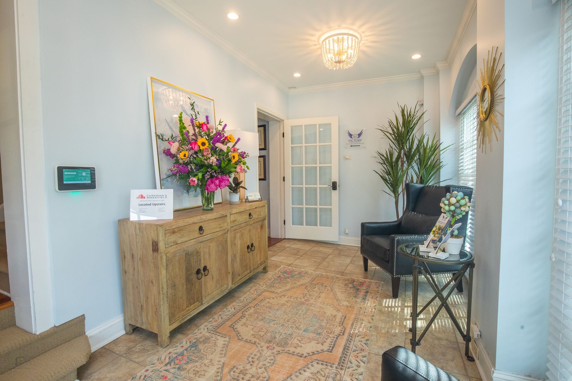Light blue entryway with ornate rug, wooden cabinet, floral arrangement, and a black armchair.