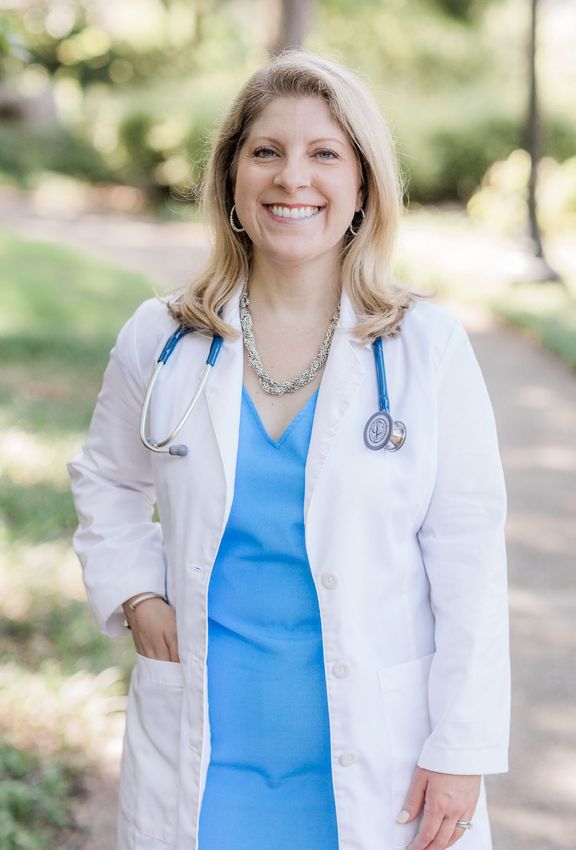 A woman in a white lab coat and blue scrubs with a stethoscope around her neck is smiling.