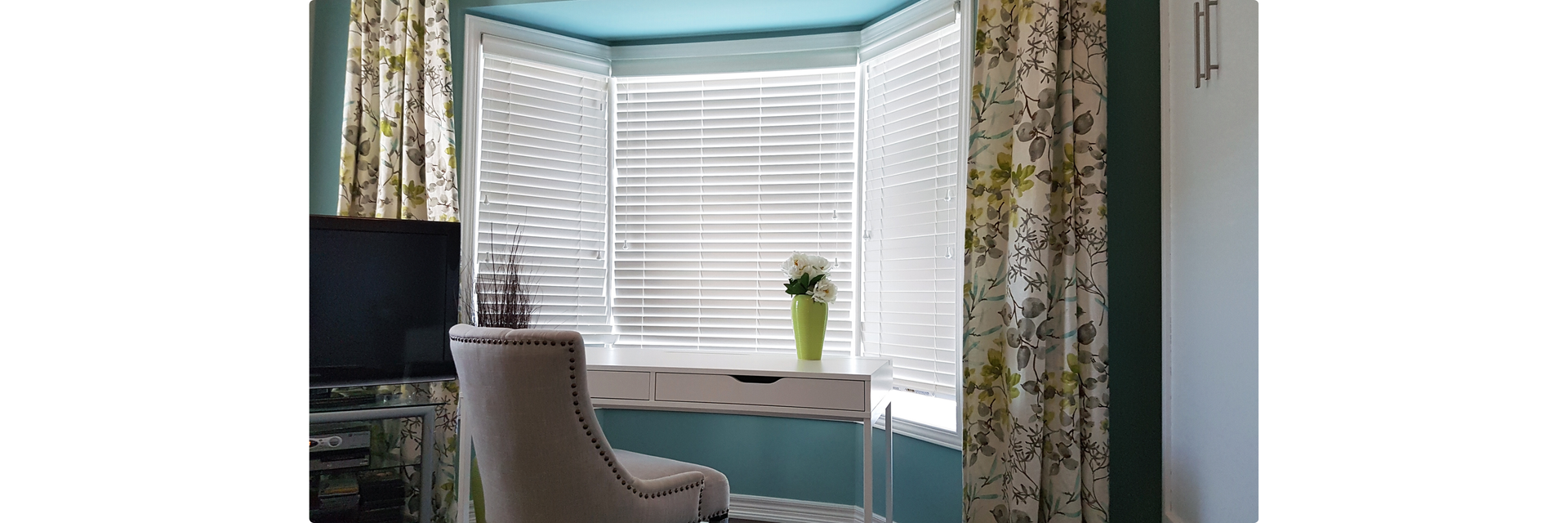 Bright bay window nook with a white desk and upholstered chair, framed by floral curtains and teal walls, creating visual balance and a sense of interior calm.