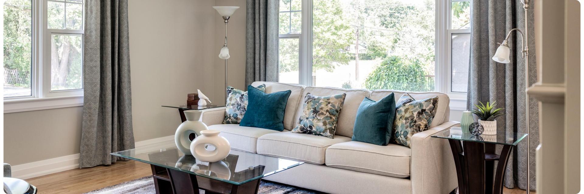 Living room with a neutral sofa, teal and floral pillows, and glass-top tables, arranged to create visual balance and interior calm through proportion and symmetry.