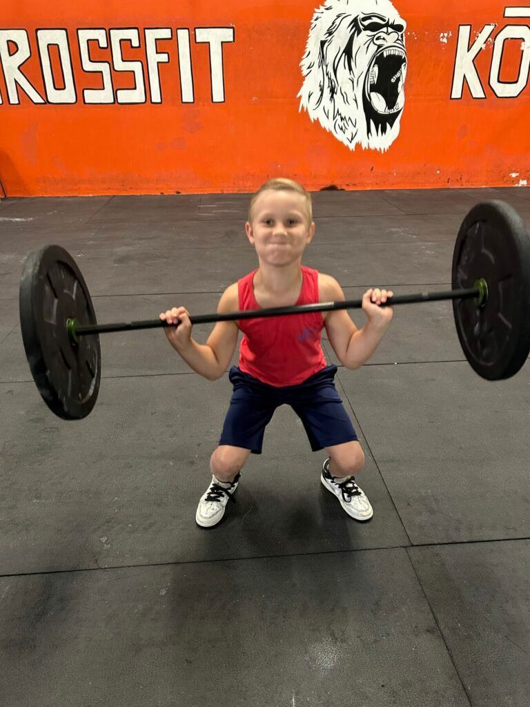 A young boy is squatting down holding a barbell in front of a sign that says crossfit