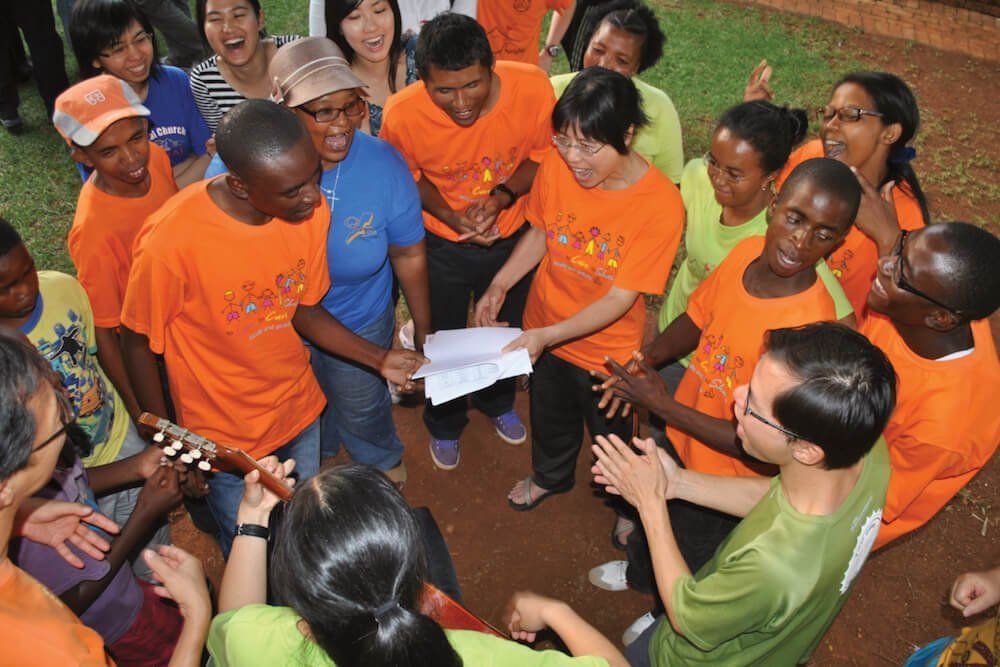 A group of people in orange shirts are gathered around a man playing a guitar