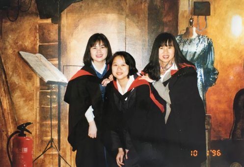 Three women in graduation gowns pose for a picture with a fire extinguisher in the background