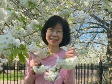 A woman in a pink sweater is standing in front of a tree with white flowers.