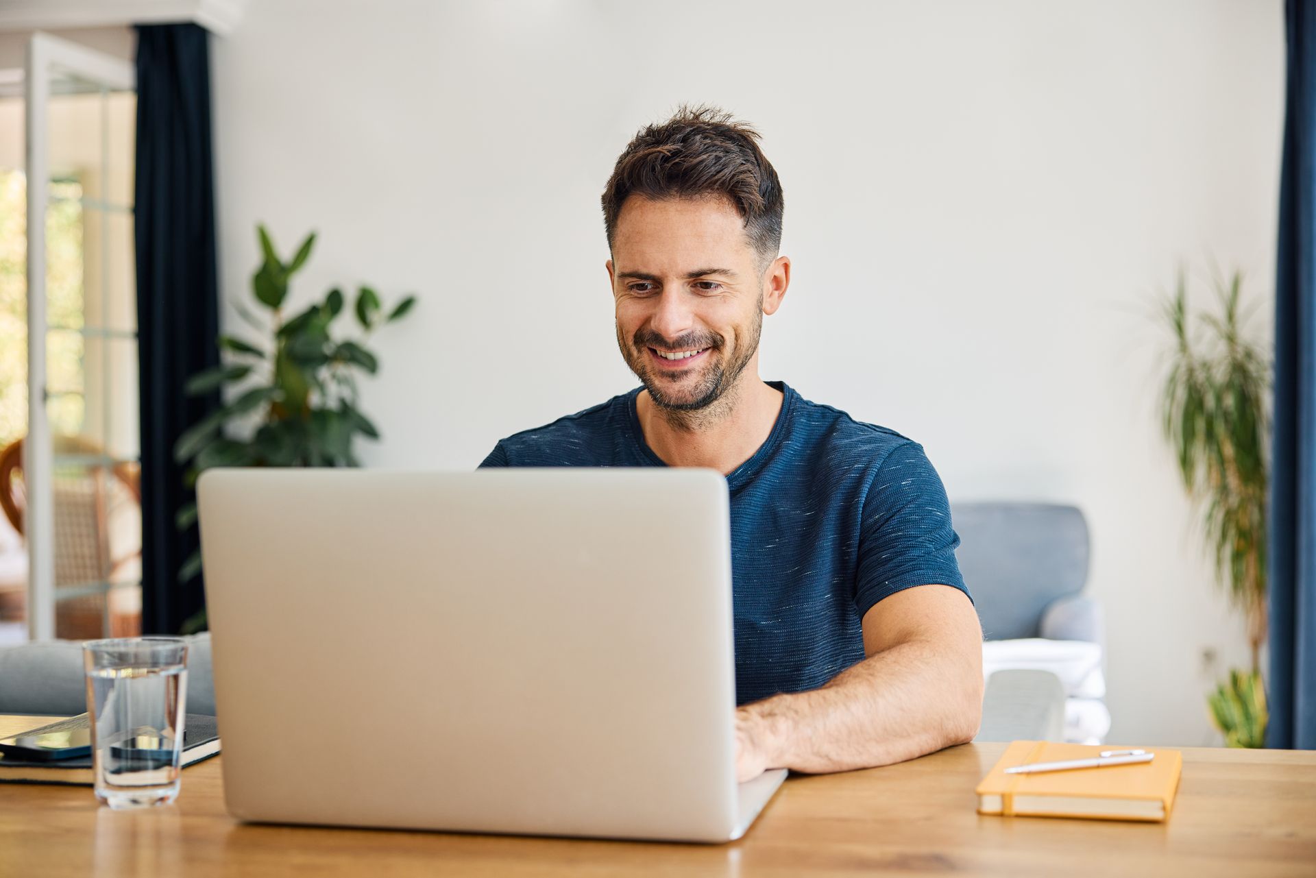 Man in blue shirt smiles while working on a laptop at a desk indoors.