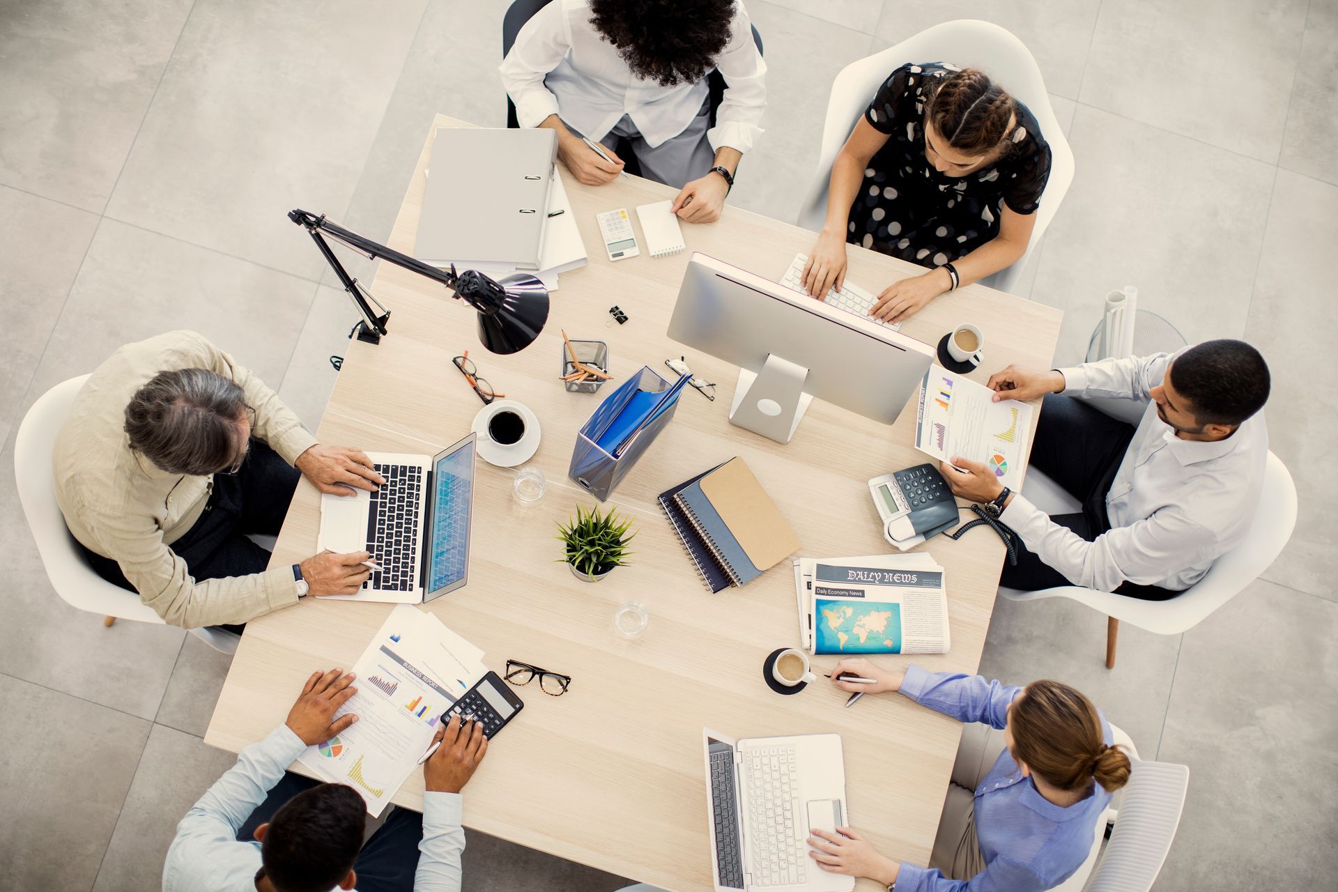 A high-angle view of six people working collaboratively around a conference table with documents, a computer, and coffee.