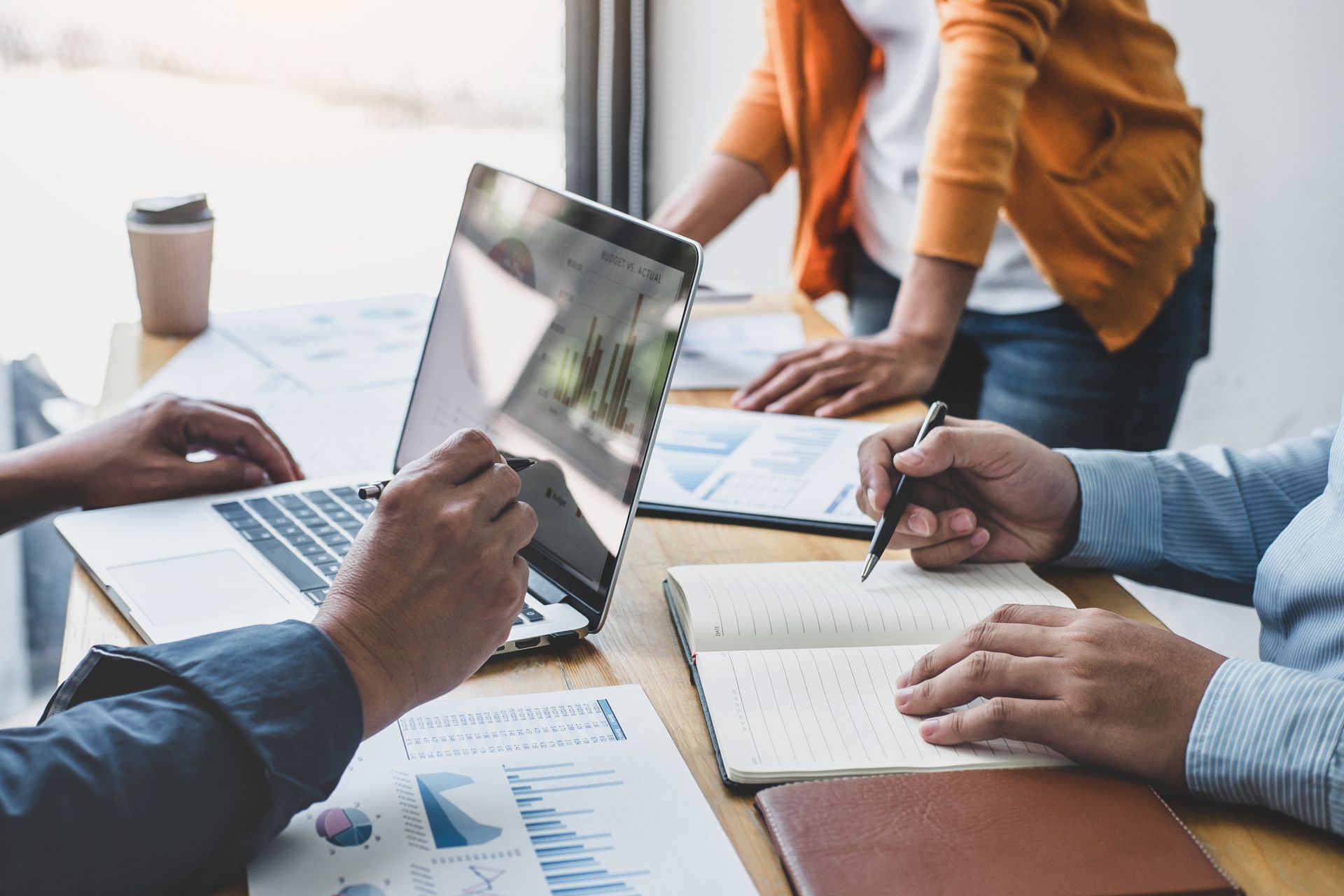 People collaborating around a table with laptop, papers, and notebook in a bright office.