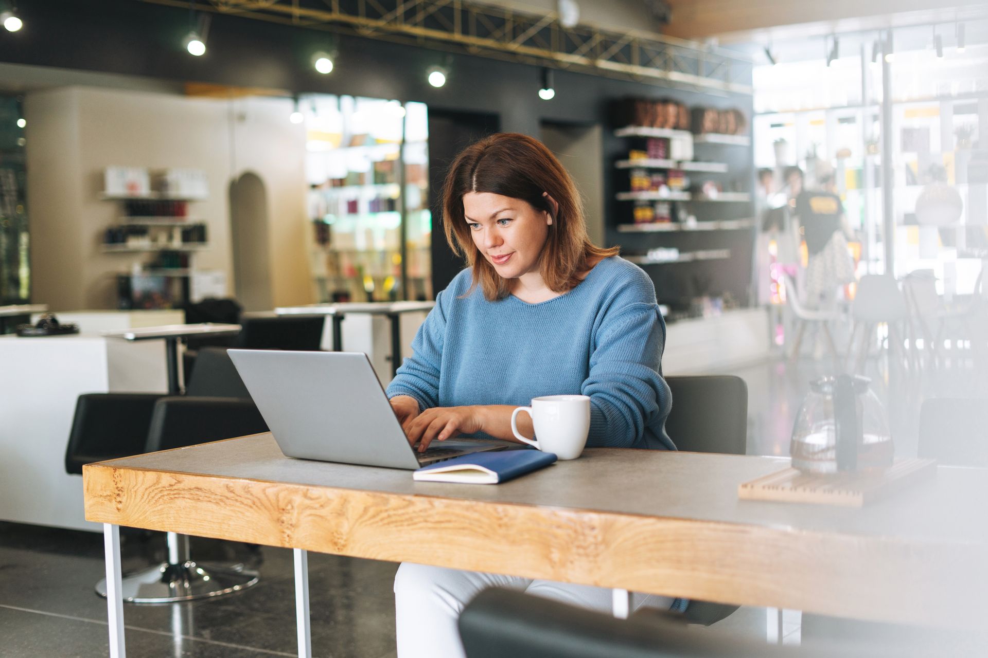 Woman working on laptop at a table in a cafe, blue sweater, coffee cup, notebook.