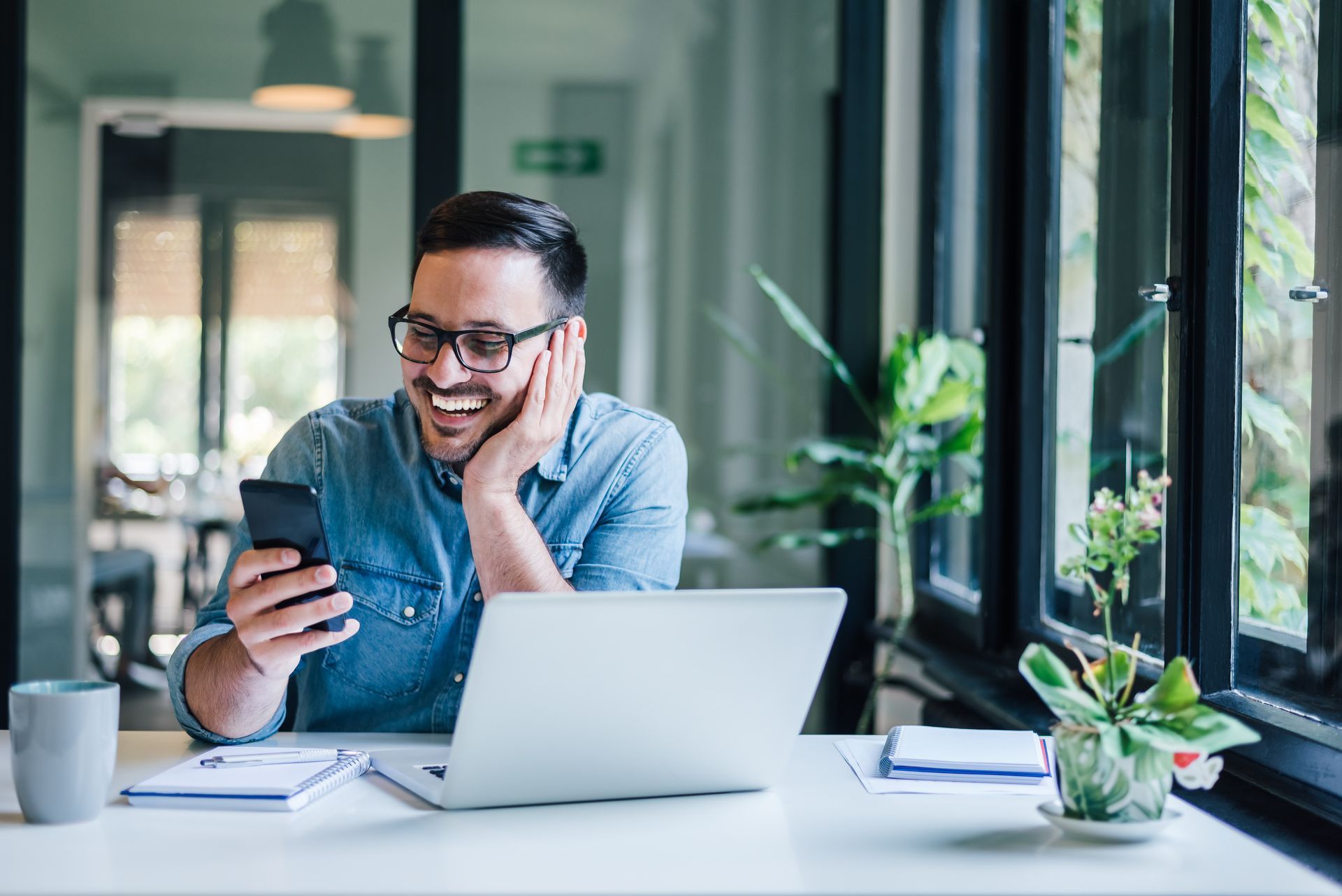 Man smiles while looking at a phone in front of an open laptop at a desk near a window.