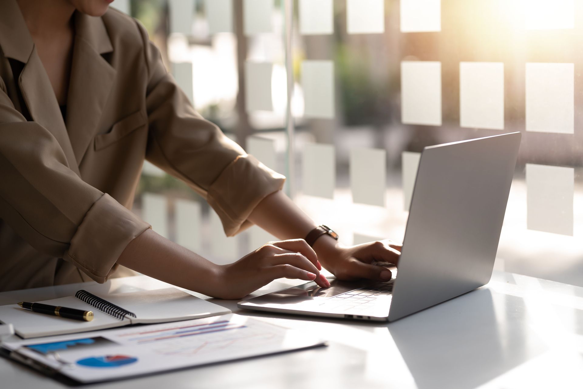 A person in a beige blazer types on a laptop at a desk with documents, next to a sunlit window with frosted glass panels.