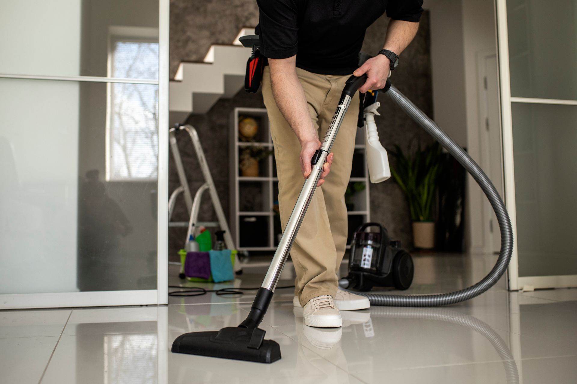 Man vacuuming a floor in a modern interior with stairs and a window in the background.