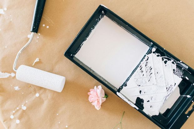Paint roller and tray with white paint, splatters, and a pink rose on a tan surface.