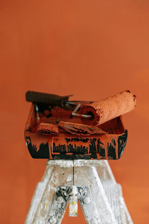A paint tray with a roller and brush sit on a small stepladder, all coated in orange paint, in front of an orange wall.