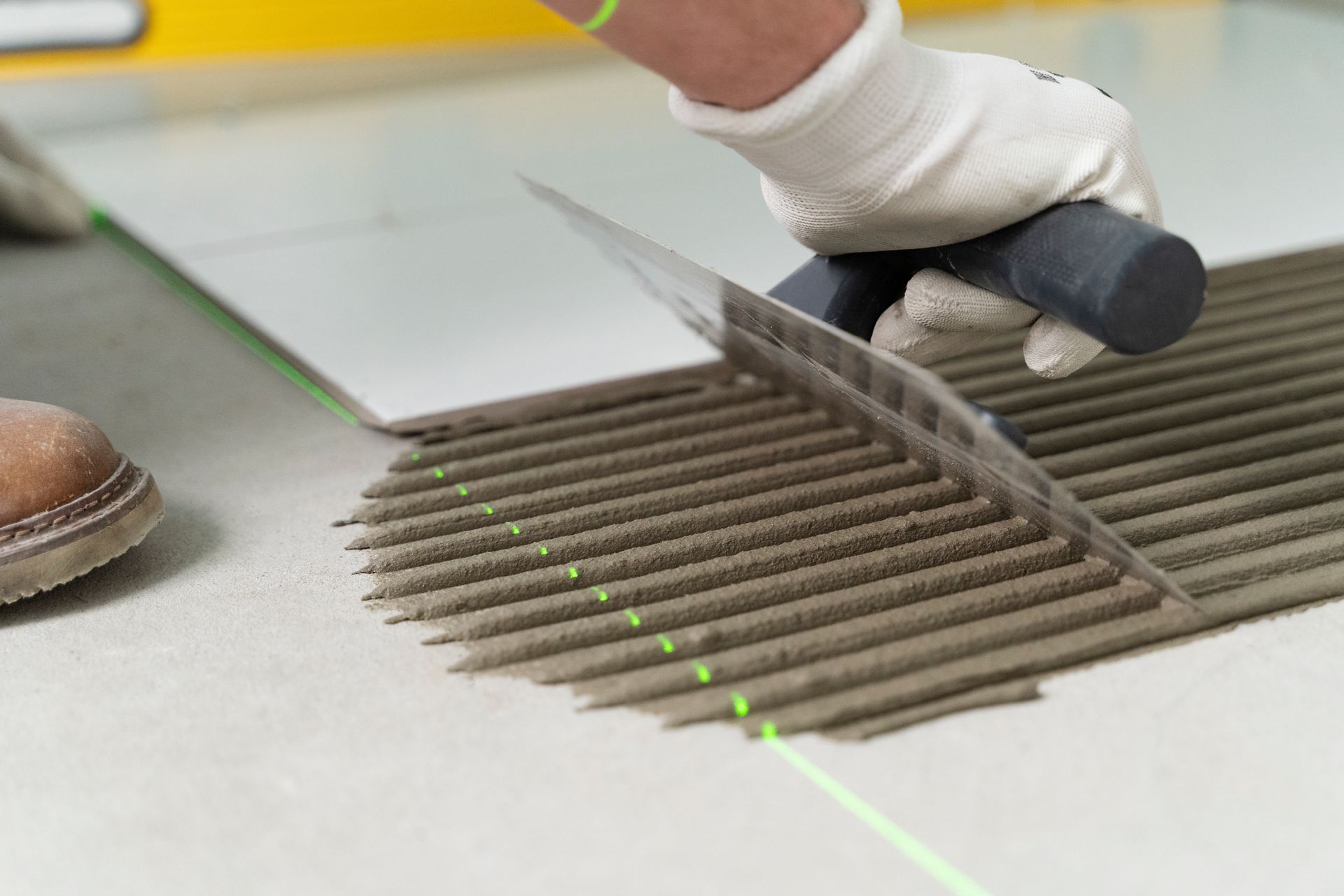 Person tiling floor with a notched trowel, creating ridges of mortar. Laser level.
