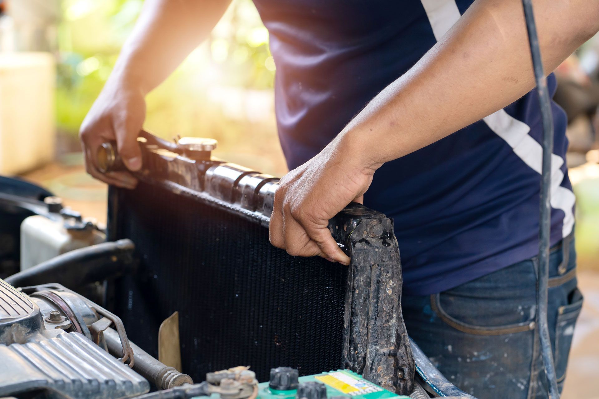 Person removing a car radiator, hands gripping the top, engine bay visible.