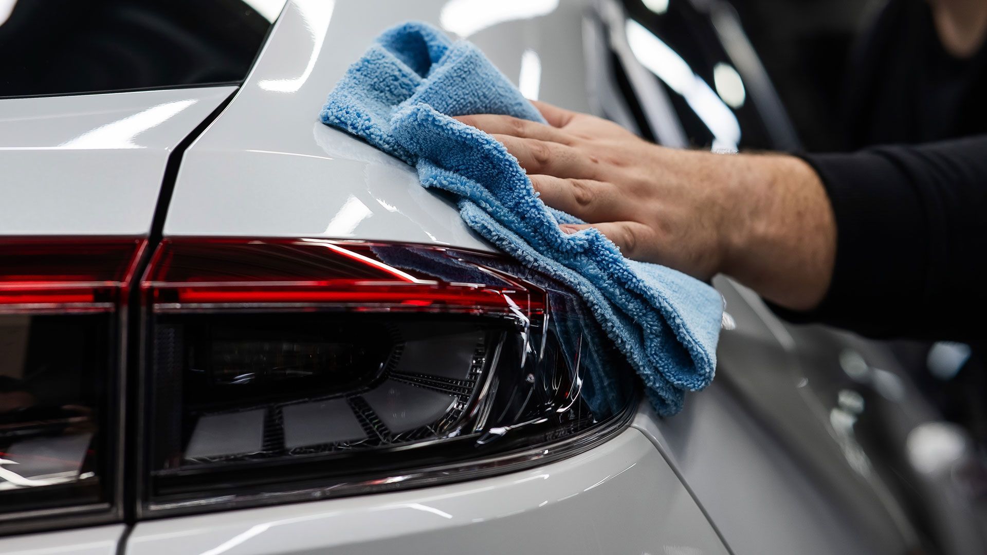 Hand wiping a blue microfiber cloth over the rear end of a white car, near a red taillight.