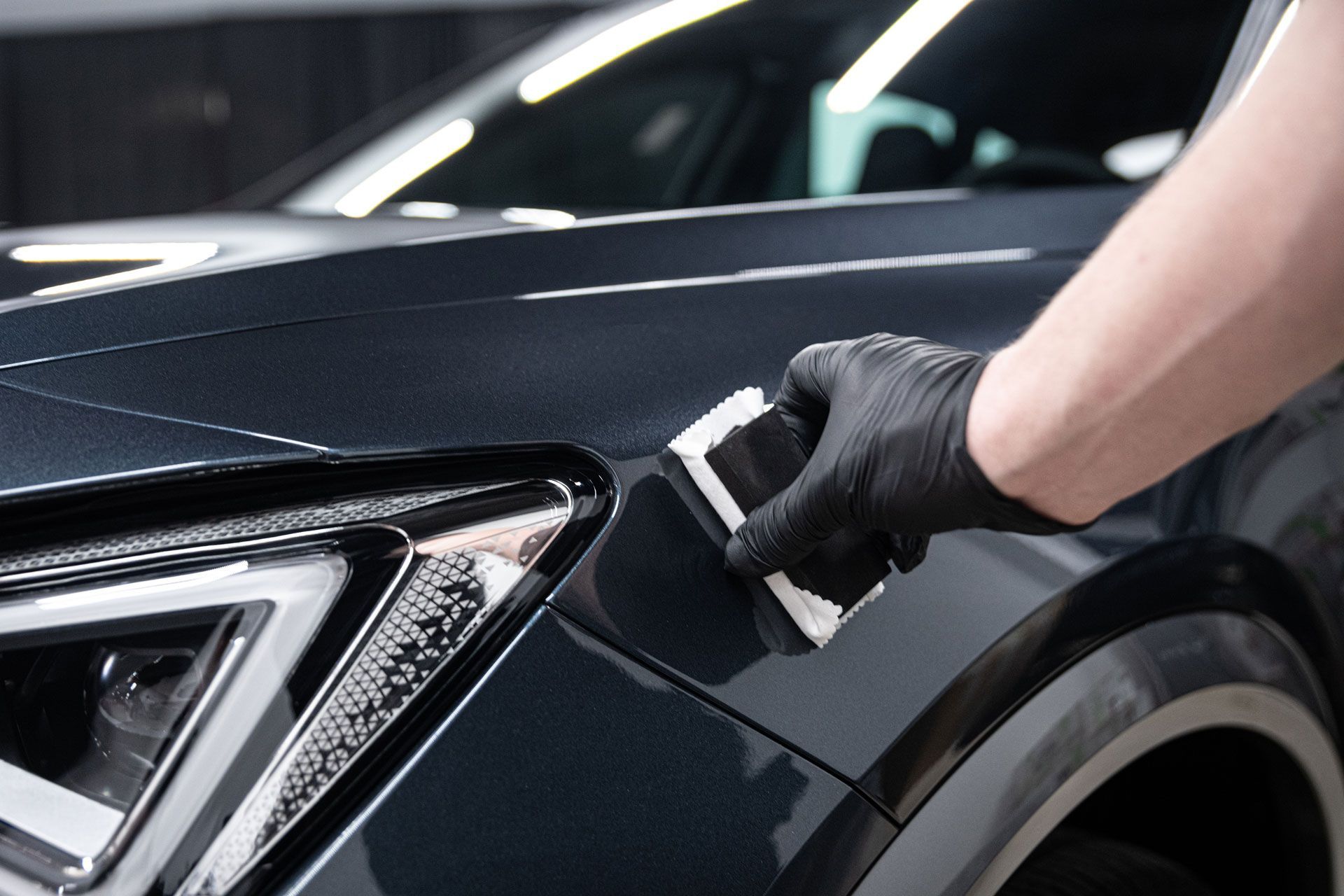 Gloved hand applying ceramic coating to a dark gray car panel in a well-lit setting.