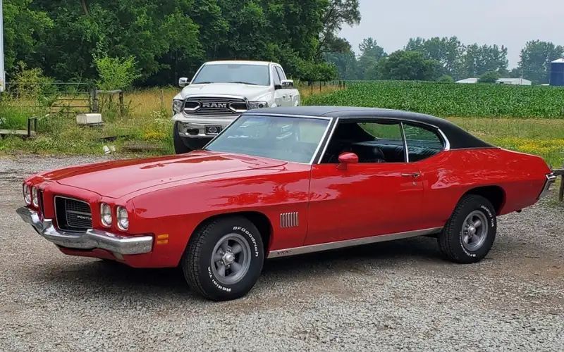 Red Pontiac LeMans with black top parked outdoors, white truck in background.