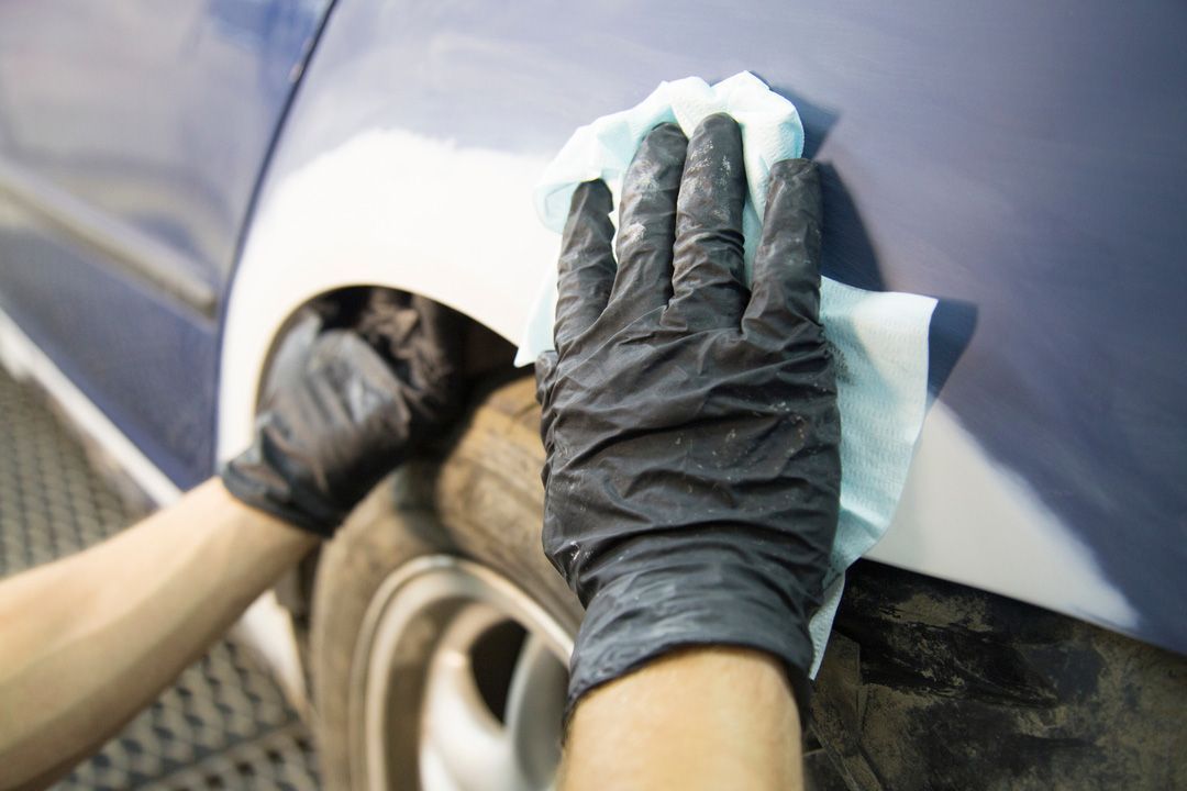 Person wearing black gloves wiping down a blue car panel with a light blue cloth.