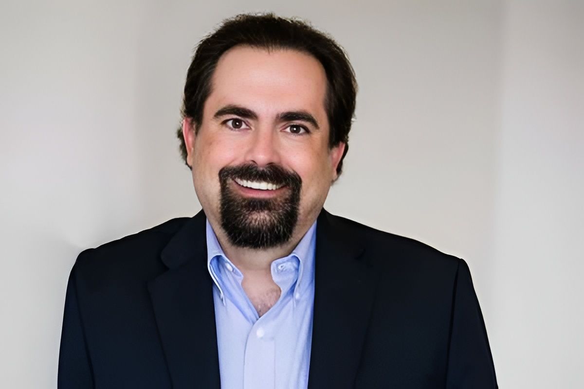 Man with dark hair and beard, wearing a blue shirt and navy blazer, smiles at the camera.