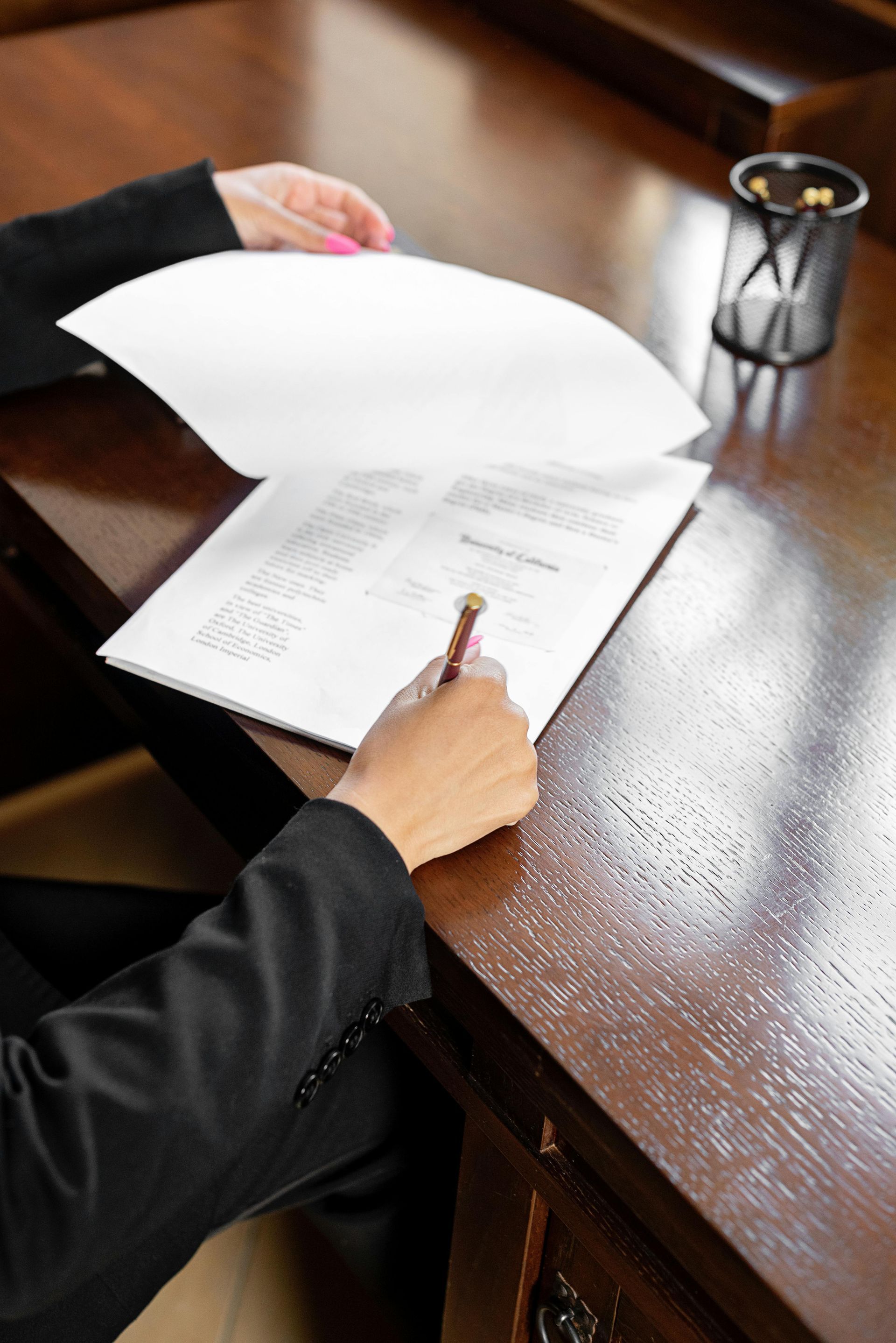 Person signing document at a wooden desk. Dark suit and pen visible.
