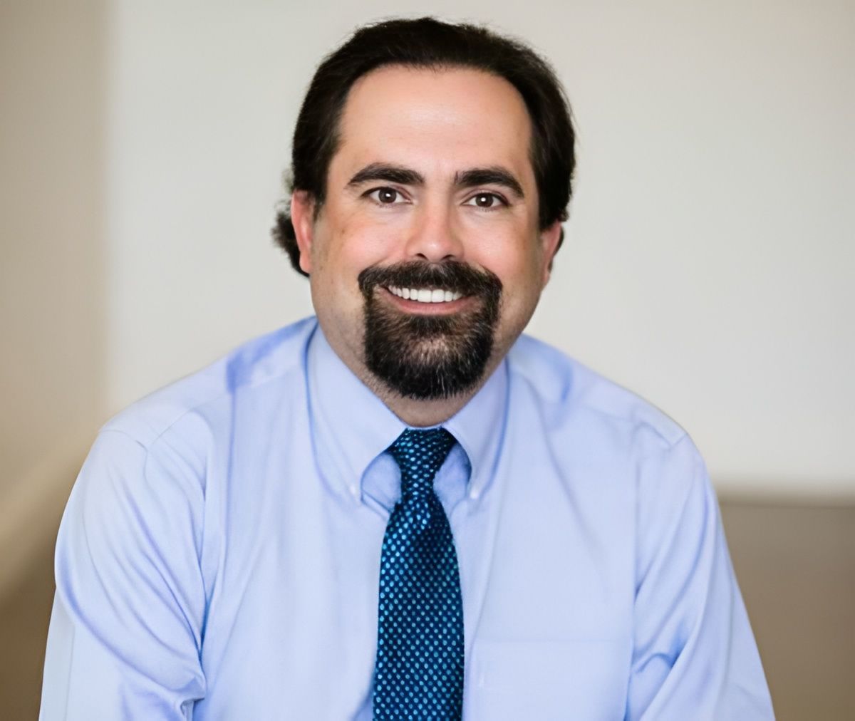 Smiling man in a blue shirt and tie with a goatee, in front of a neutral background.