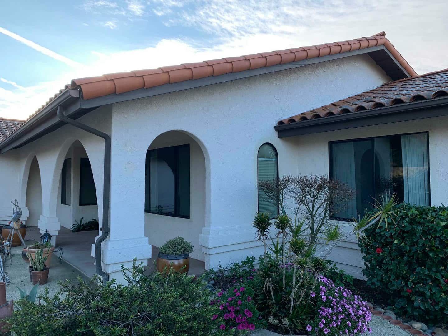 A white house with a red tile roof is surrounded by flowers and bushes.