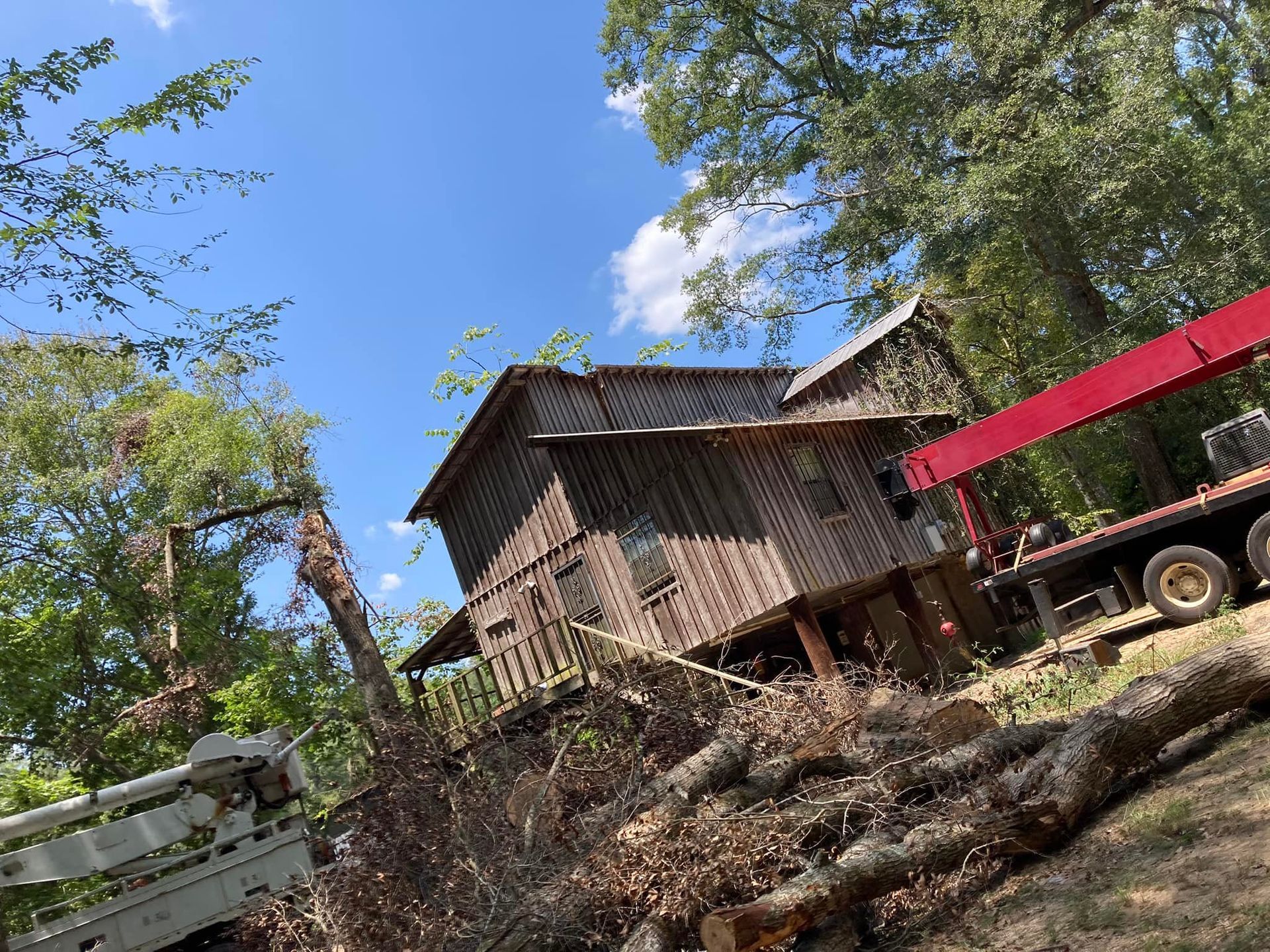 Old wooden house being worked on by a crane; set amongst trees under a blue sky.