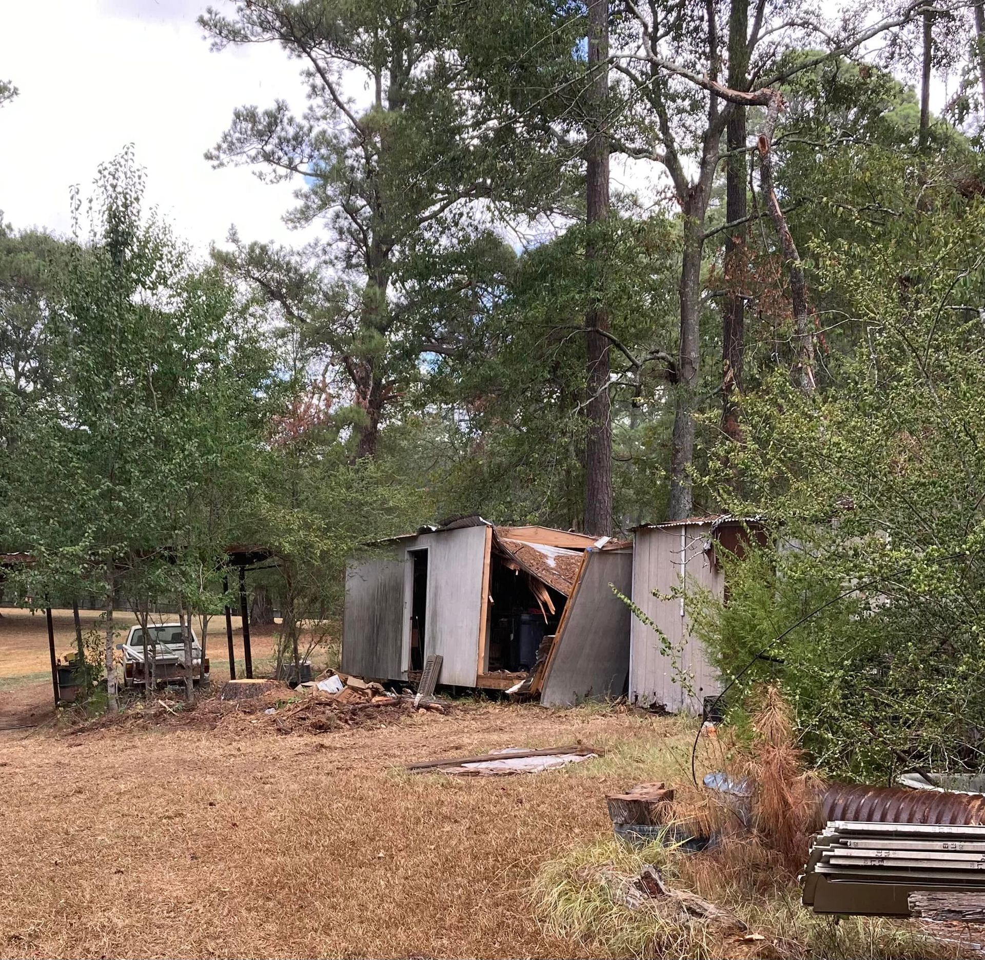 Damaged shed in a rural area, with debris and trees. Overcast sky and brown grass.