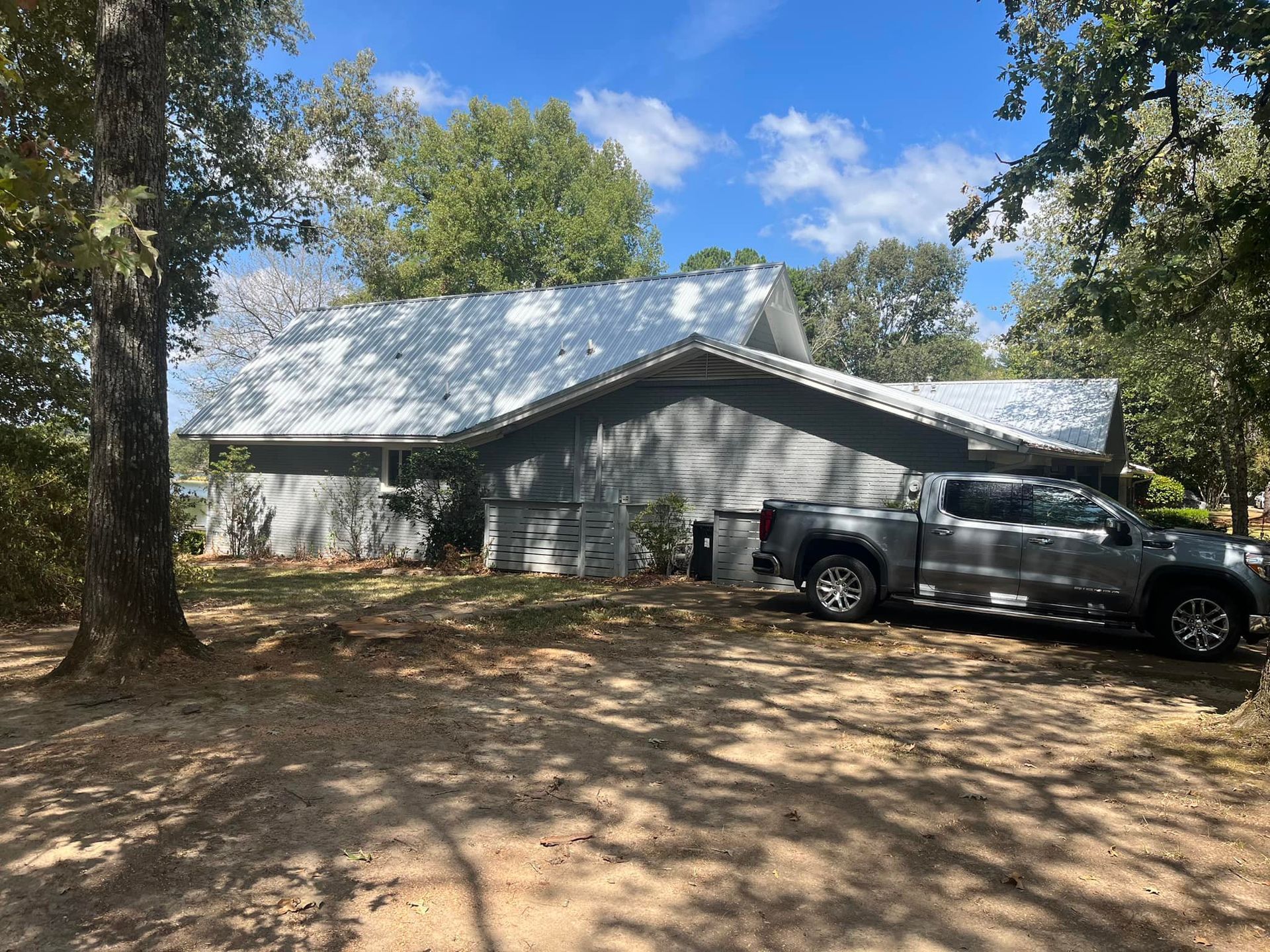 Gray house with a shiny metal roof, parked gray truck, surrounded by trees on a sunny day.