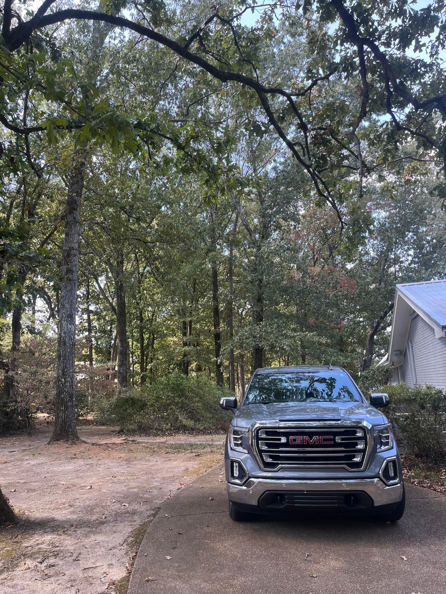 Gray GMC truck parked on a curved driveway in a wooded area with a white house in the background.