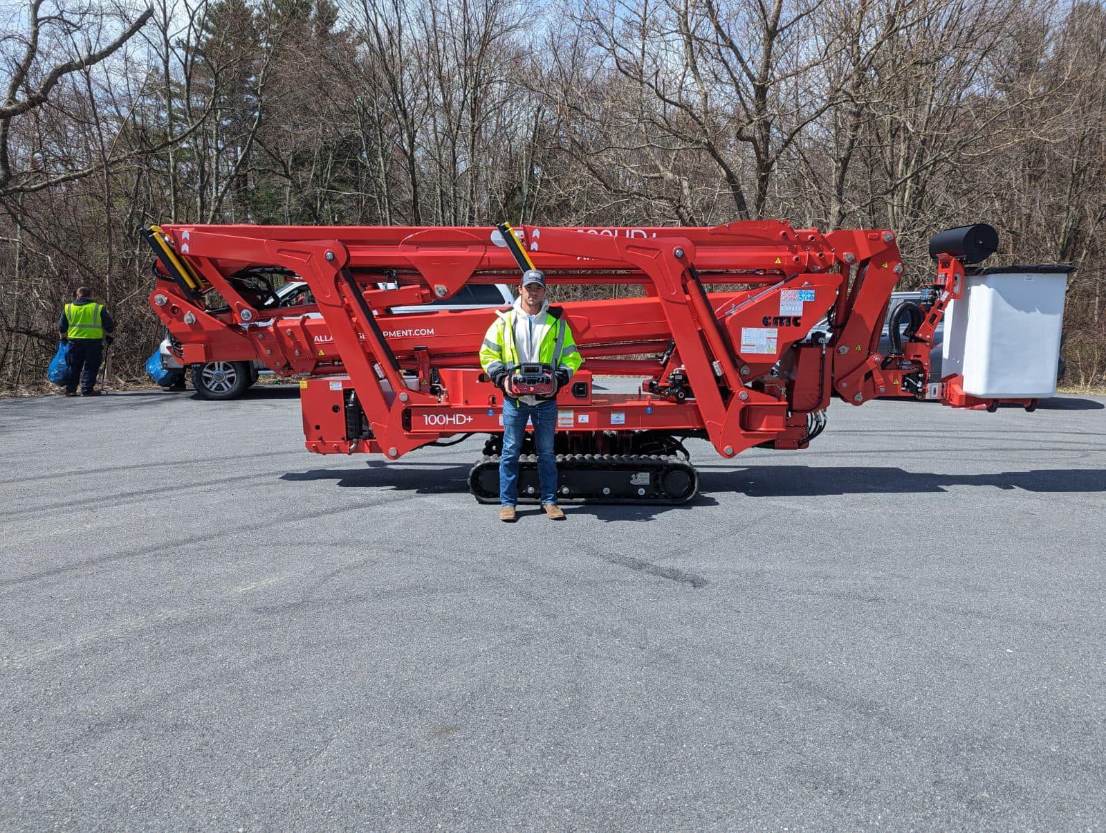 Man operates a large red spider lift on a paved lot; trees in the background.