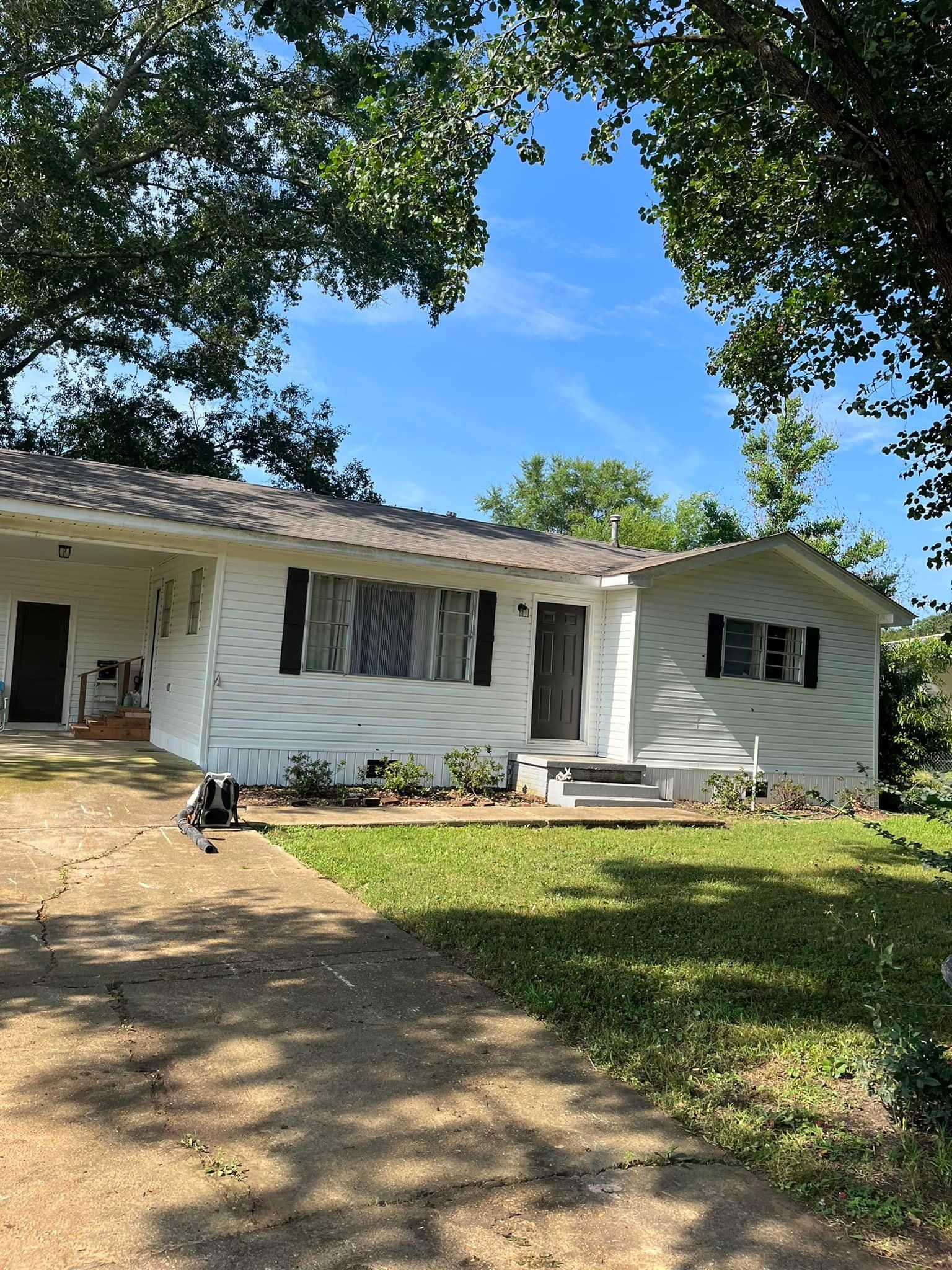 White house with black shutters, a porch, and green lawn under a blue sky, framed by tree branches.