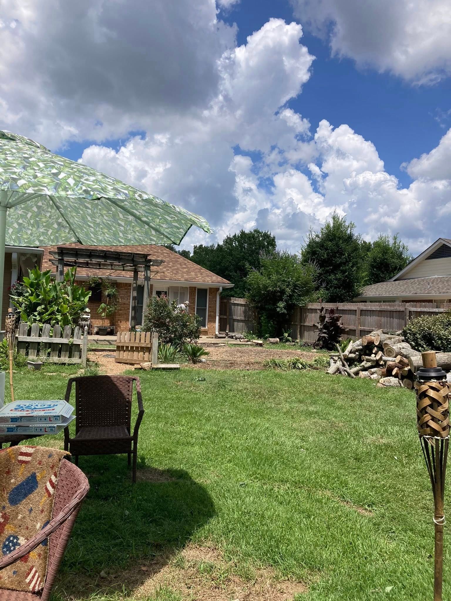 Backyard scene: green grass, brown chair, patterned umbrella, small house, blue sky with clouds.