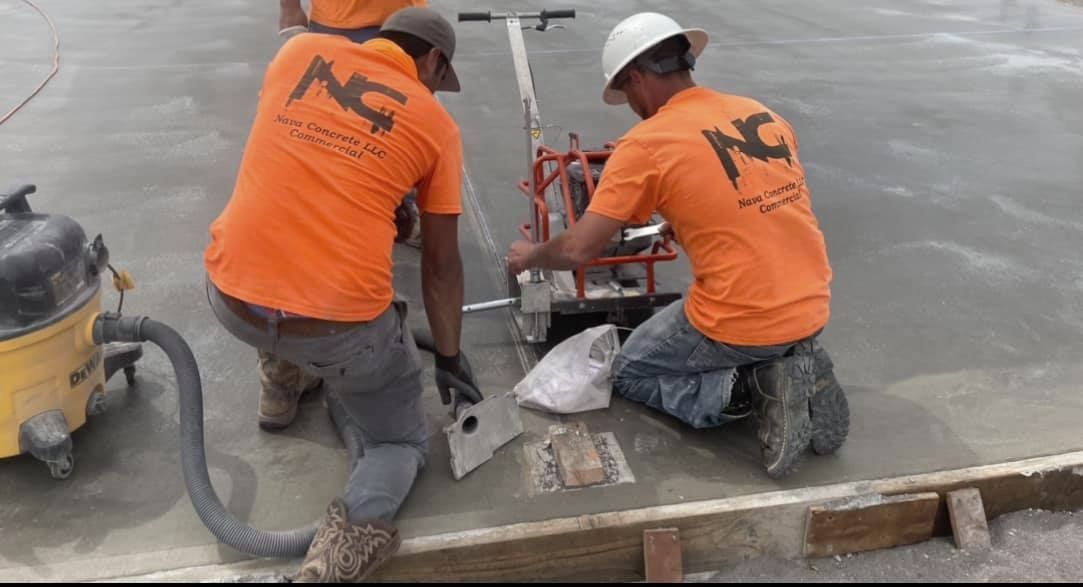 Two construction workers wearing orange shirts are working on a concrete floor.