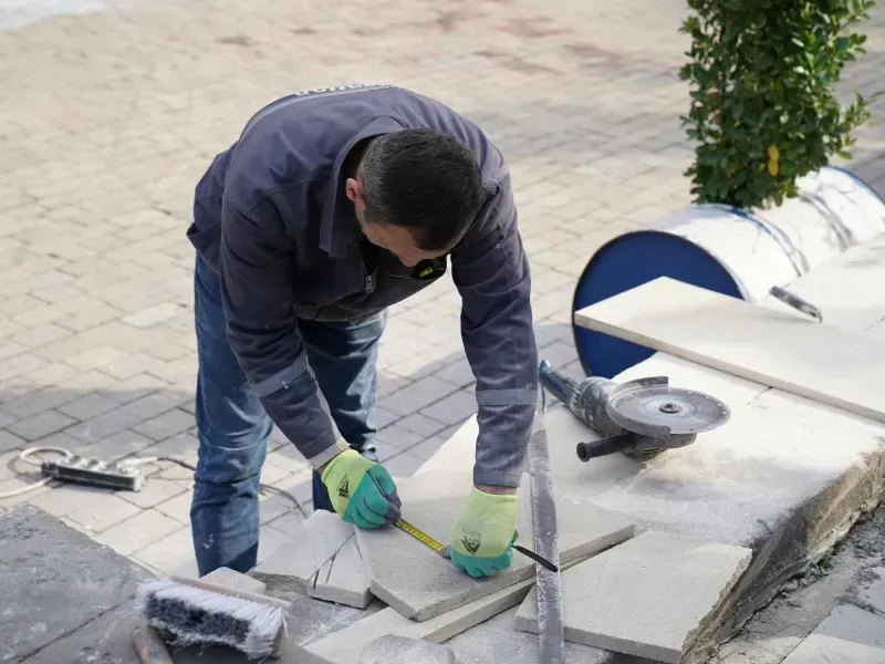 Man measuring a tile with a tape measure, preparing to cut it with a grinder, outdoors.